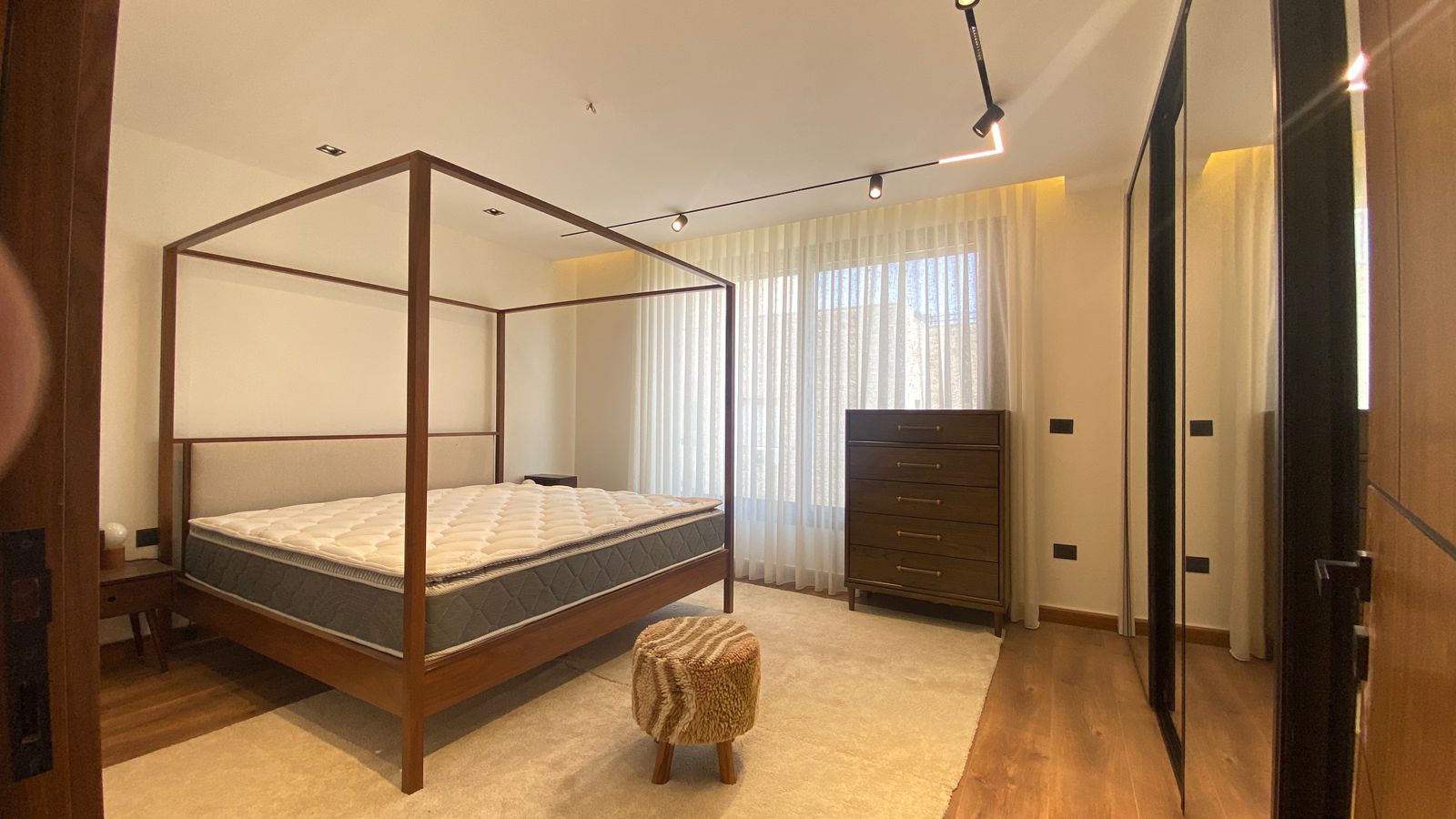 c3adc2bc-6add-47bf-b0af-5eccf5bf65c8 Wood four-poster bed with tufted headboard in a bright modern bedroom, with a dresser and large window with sheer curtains.