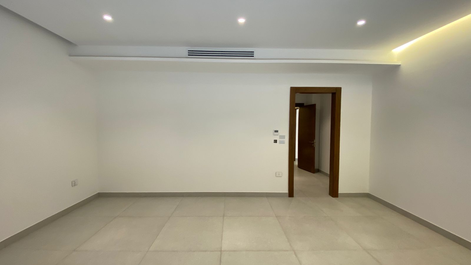 c448ed10-ba87-4b73-bf2a-a651d636dc6b Empty modern room with white walls, light gray tiled floor, and a wooden doorway opening to another room; ceiling recessed lights and a right-edge LED strip lighting.