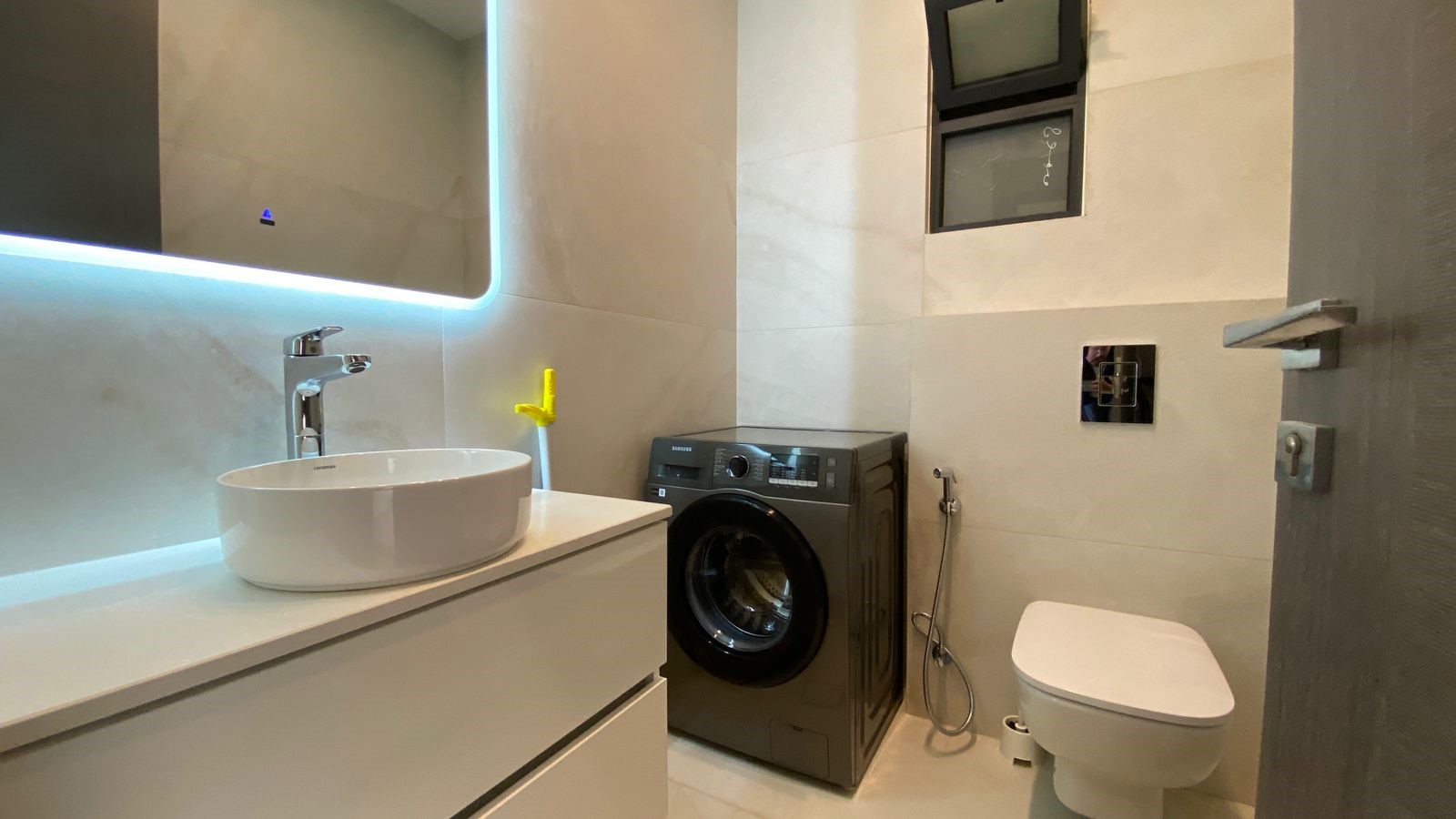 c481284c-afc0-40be-aeef-7e28c54dd1da Modern bathroom with a white vanity, round basin, and backlit rectangular mirror; a gray washing machine sits against the wall, near a wall-mounted toilet with a bidet hose nearby.