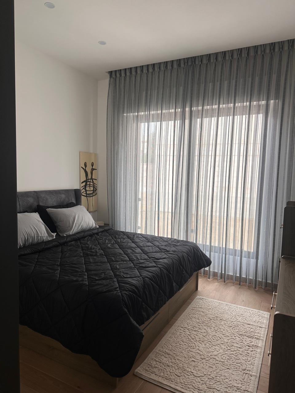 c4f60dd0-898a-46a5-a041-dc0f14c8448c Bedroom with a low wooden bed, black quilted cover, white pillows, and sheer gray curtains by a large window.