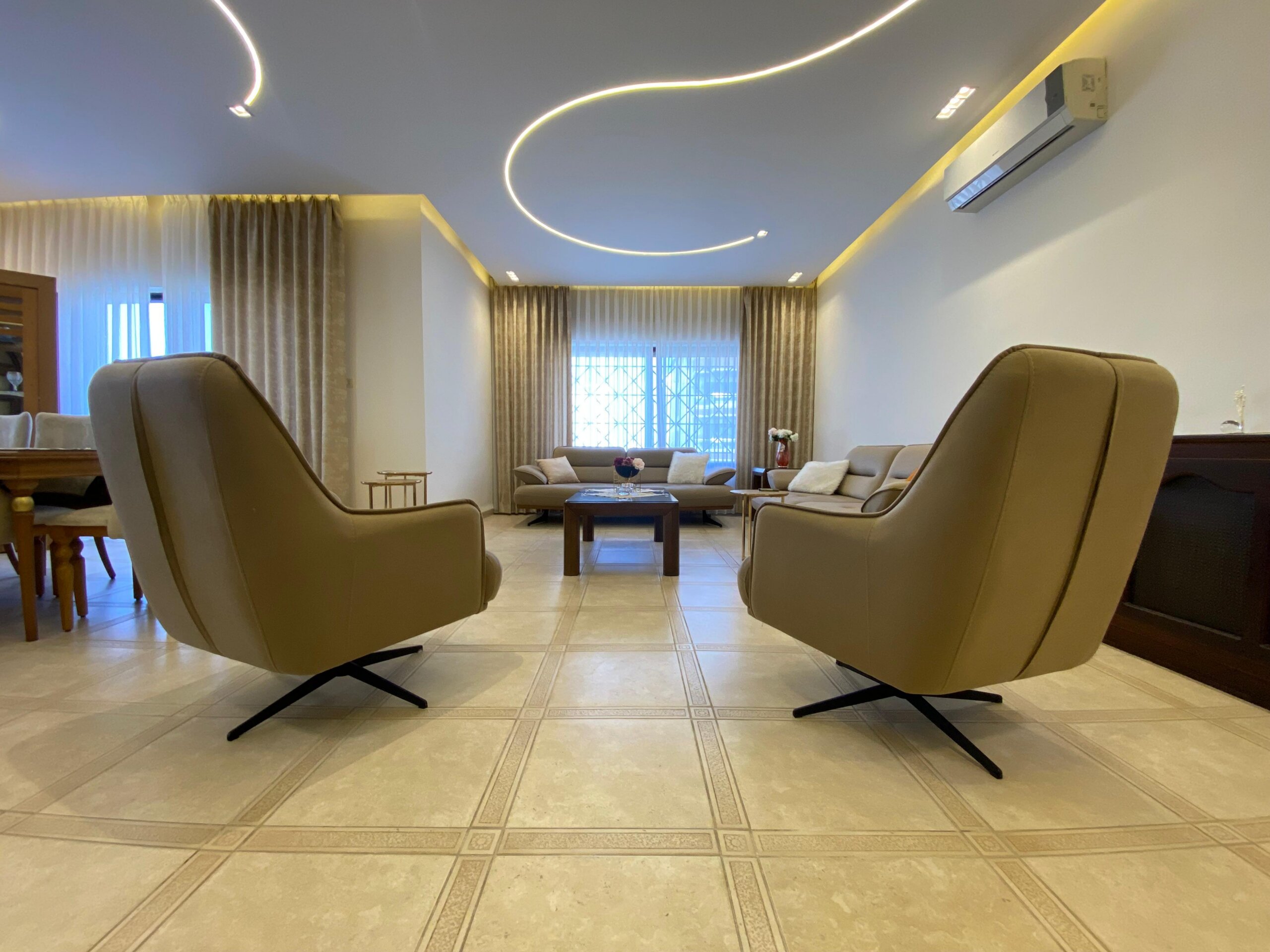 c5076687-871c-4ce8-94b2-551b0680d714 Modern living room with beige lounge chairs facing a central coffee table, sofas in the background, large windows with sheer curtains, and curved neon ceiling lights.