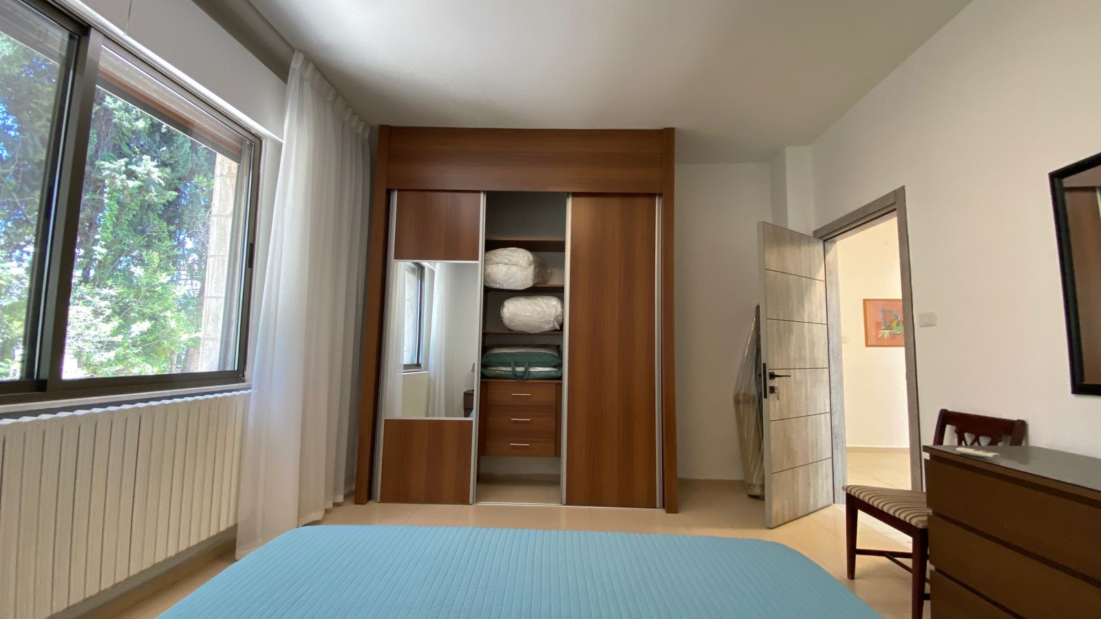 c5ad10d1-43f4-4323-9b7e-11d58feaafbc Bedroom interior with a large window on the left, sheer curtains, and a built-in wooden wardrobe showing folded linens and drawers opposite the bed with a blue cover.