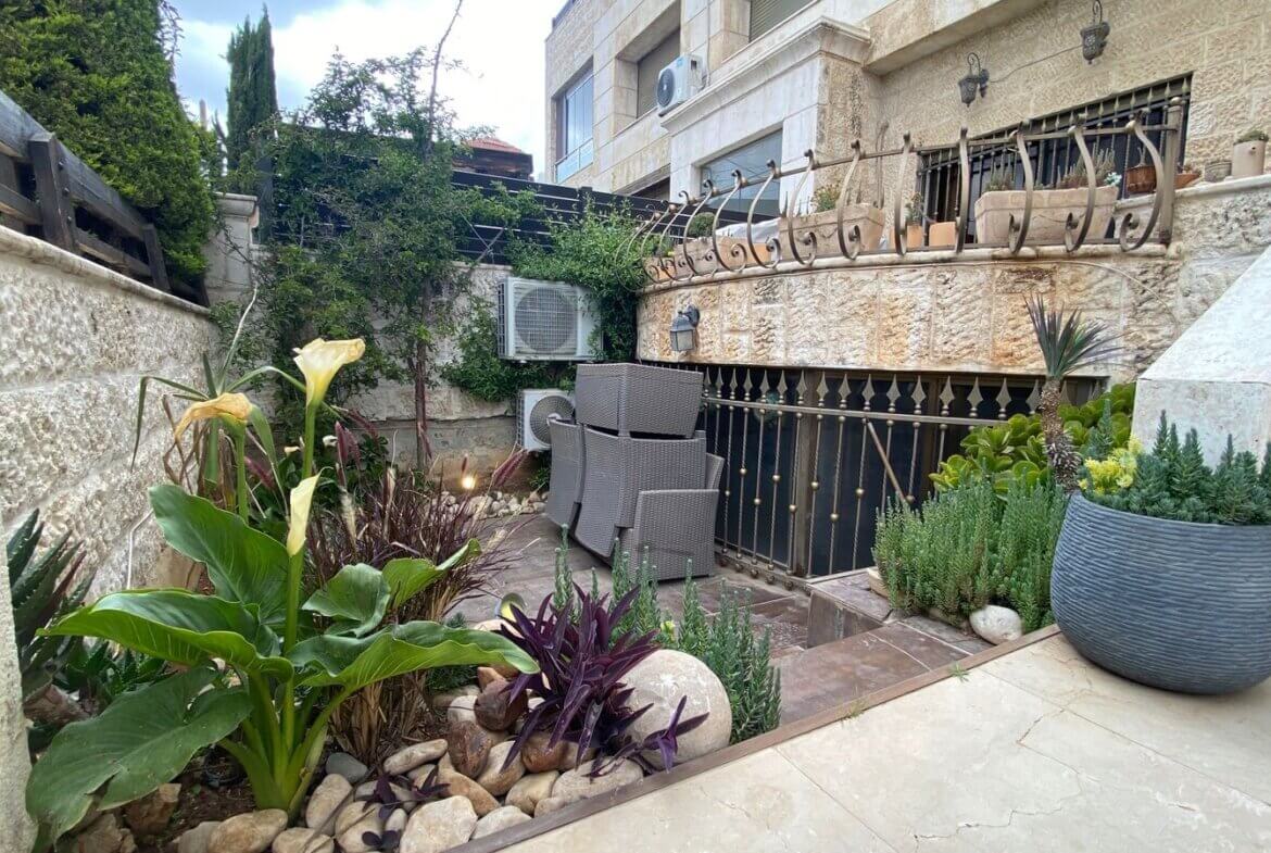 c603bd27-dbb7-4886-90b5-f10ed2f2b171 Stone-walled courtyard with stacked outdoor wicker chairs, potted plants, and a stair railing; leafy plants and rocks line the garden bed.