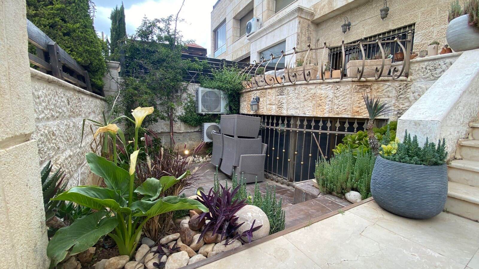 c603bd27-dbb7-4886-90b5-f10ed2f2b171 Stone-walled courtyard with stacked outdoor wicker chairs, potted plants, and a stair railing; leafy plants and rocks line the garden bed.