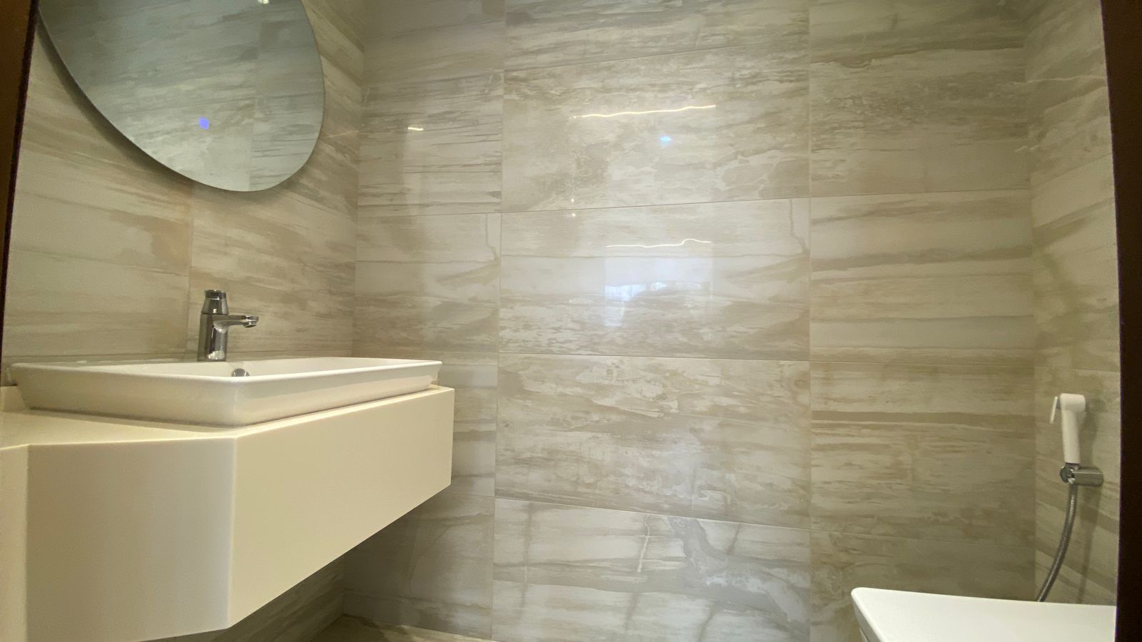 c6631e2f-b9c7-45ce-8cc6-cf601c401439 Modern bathroom with a wall-mounted white sink, chrome faucet, round mirror, and marble tile walls.