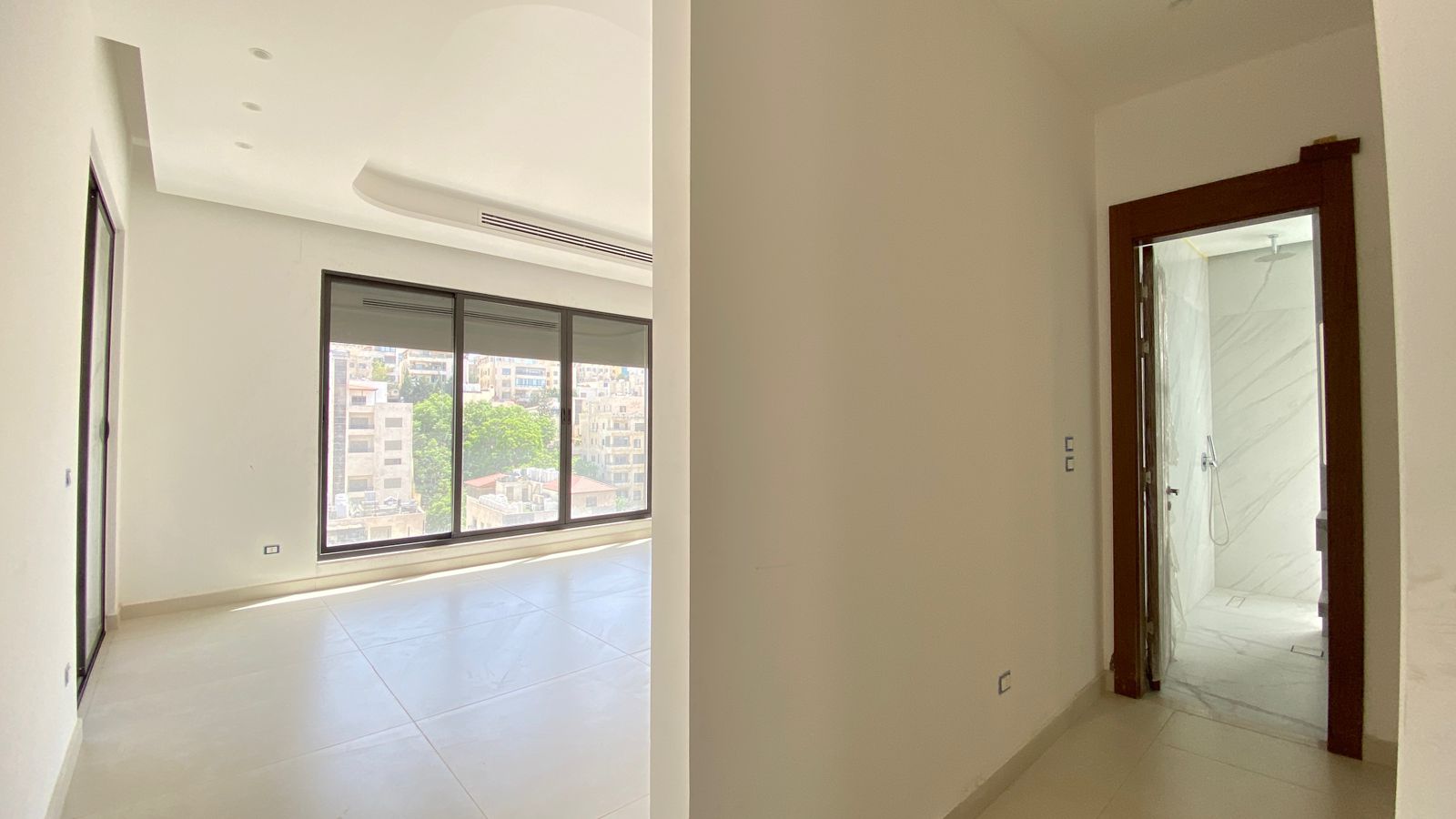 c7792481-a435-4063-90f0-2191ea01ffc3 Bright empty living room with large glass doors offering a city view, white walls and glossy tile floor; a marble bathroom is visible through a wooden-framed doorway on the right.
