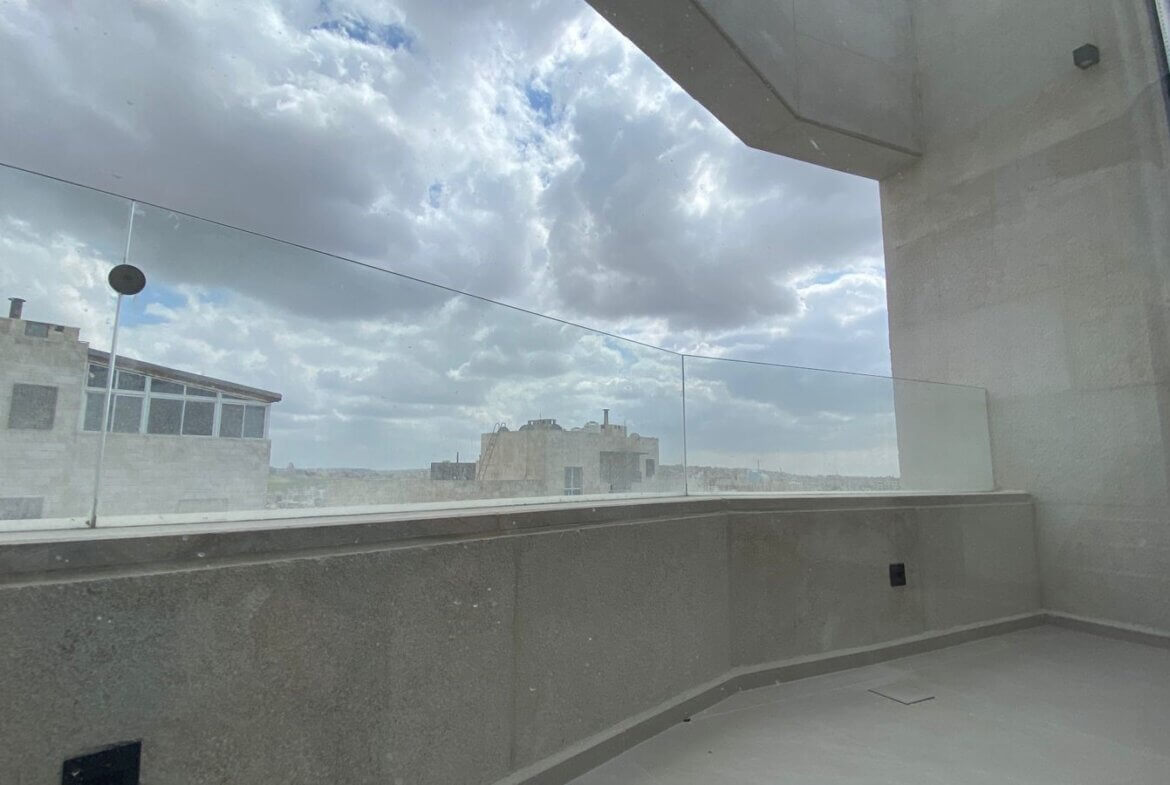 Rooftop balcony with a glass railing, grey stone walls, and a cloudy sky over distant buildings.