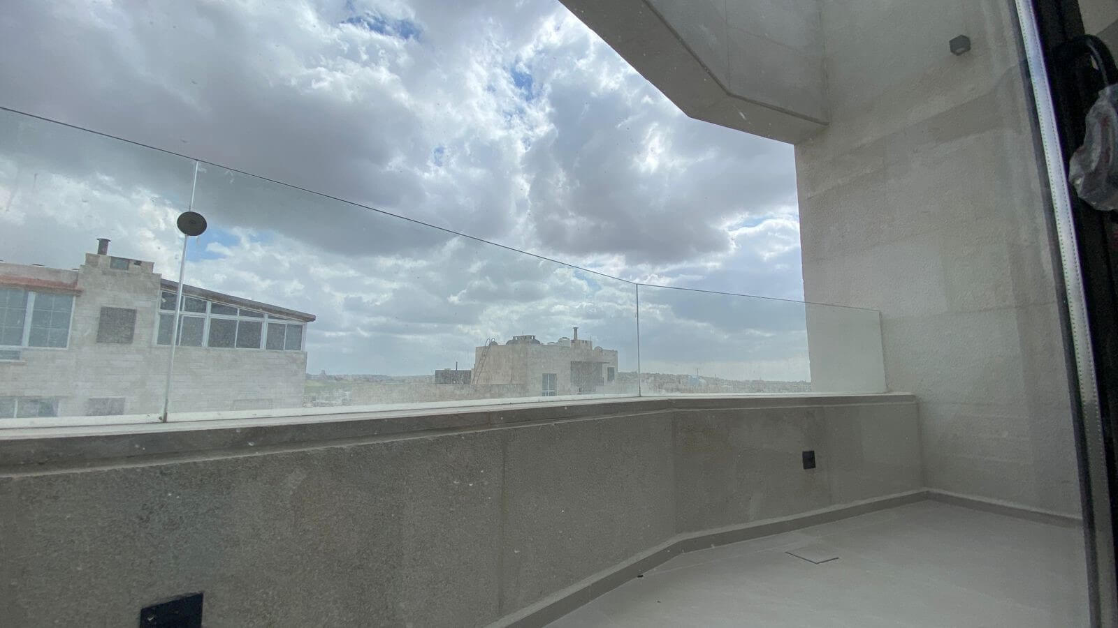 Rooftop balcony with a glass railing, grey stone walls, and a cloudy sky over distant buildings.