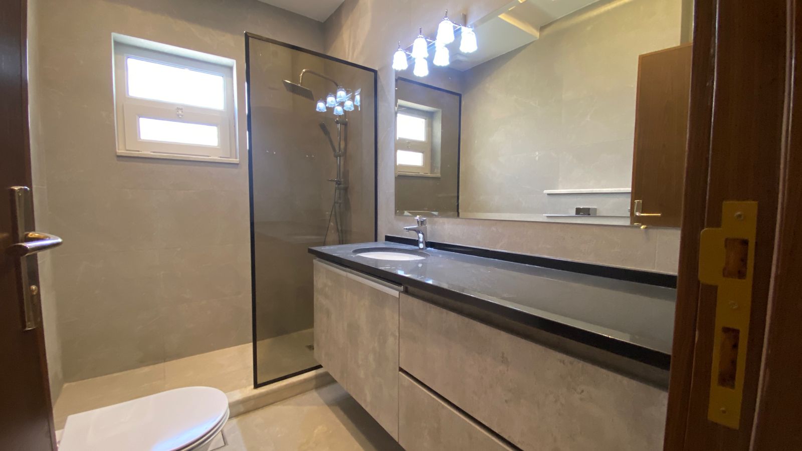 c8233b3b-4098-407f-b750-16e52a03a2bf Modern bathroom with glass shower enclosure, grey tiled walls, and a long grey vanity with a black countertop and integrated sink.