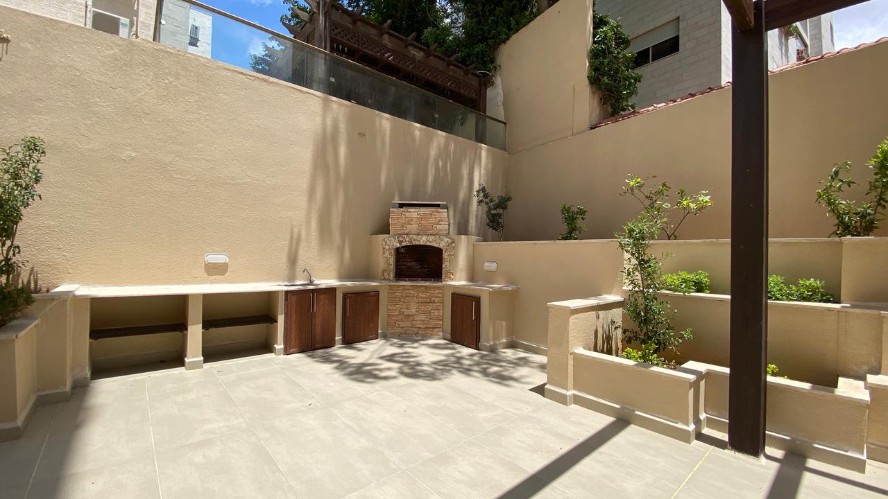 c82b57c6-b521-4467-a85f-ac91e0cf8055 Outdoor backyard kitchen with a built-in brick oven, stone facade, and beige countertops along a tan wall area.