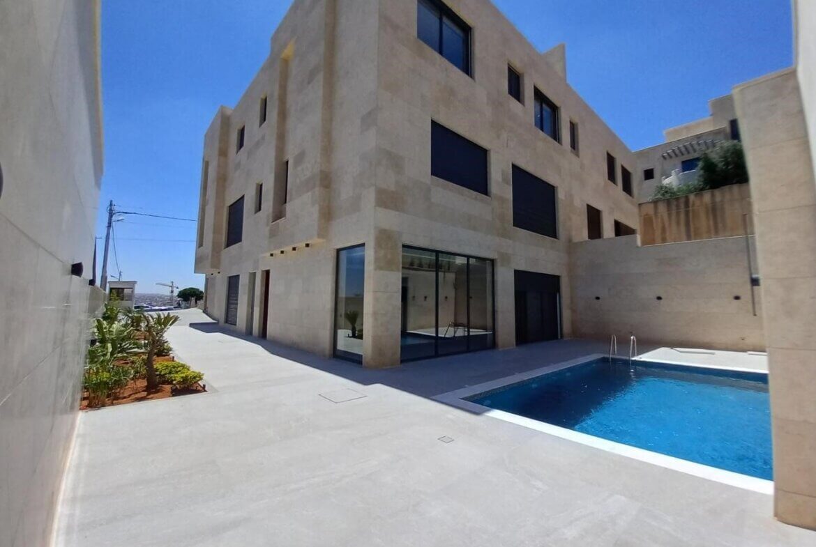 Modern beige stone building with large glass doors beside a blue rectangular pool in a fenced courtyard.