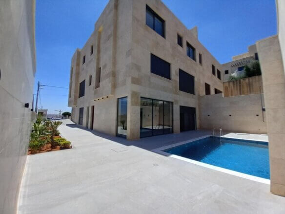 Modern beige stone building with large glass doors beside a blue rectangular pool in a fenced courtyard.