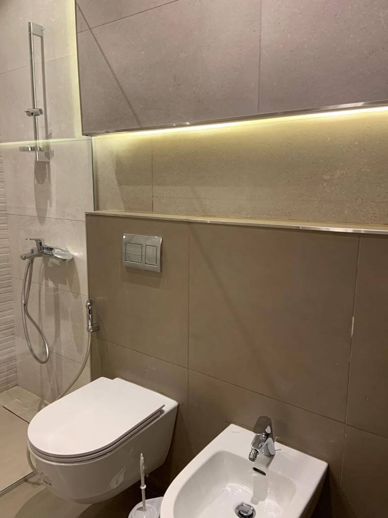 c97fd426-133a-4259-9903-fba830051843 Modern bathroom with wall-mounted toilet, bidet, and glass shower enclosure under warm LED lighting