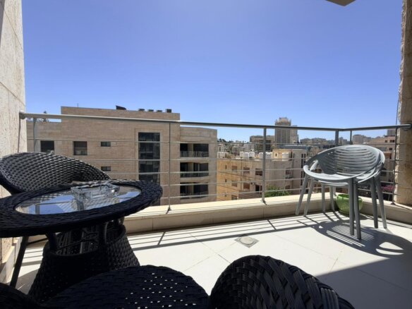 A sunny urban balcony with black wicker chairs and a glass-topped table, overlooking beige buildings and a clear blue sky.