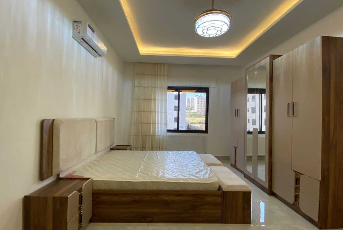 Modern bedroom with a low wooden bed, beige upholstered headboard, and matching nightstands, plus a large mirrored wardrobe.