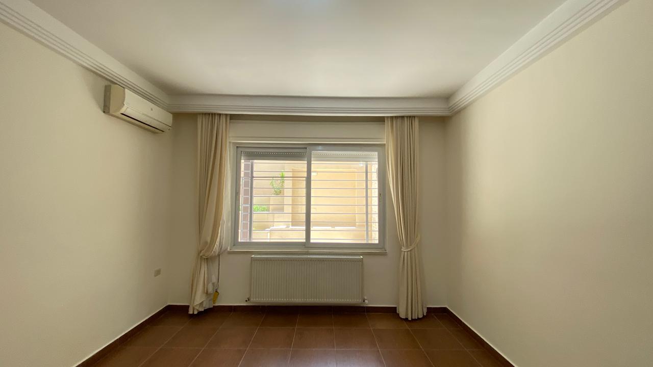 ca4fd266-0c48-450a-bfaf-ce47af0322d0 Empty rectangular room with a large window centered on the far wall, cream curtains, radiator beneath the window, and a wall-mounted air conditioner on the left.