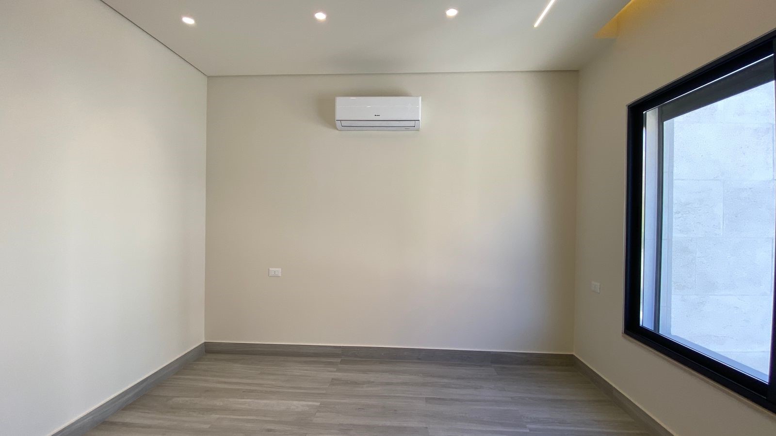 ca7bf5f1-fb0a-4a58-889f-e83179338808 Empty modern room with beige walls, gray wood-like flooring, a wall-mounted air conditioner, and a large right-side window.