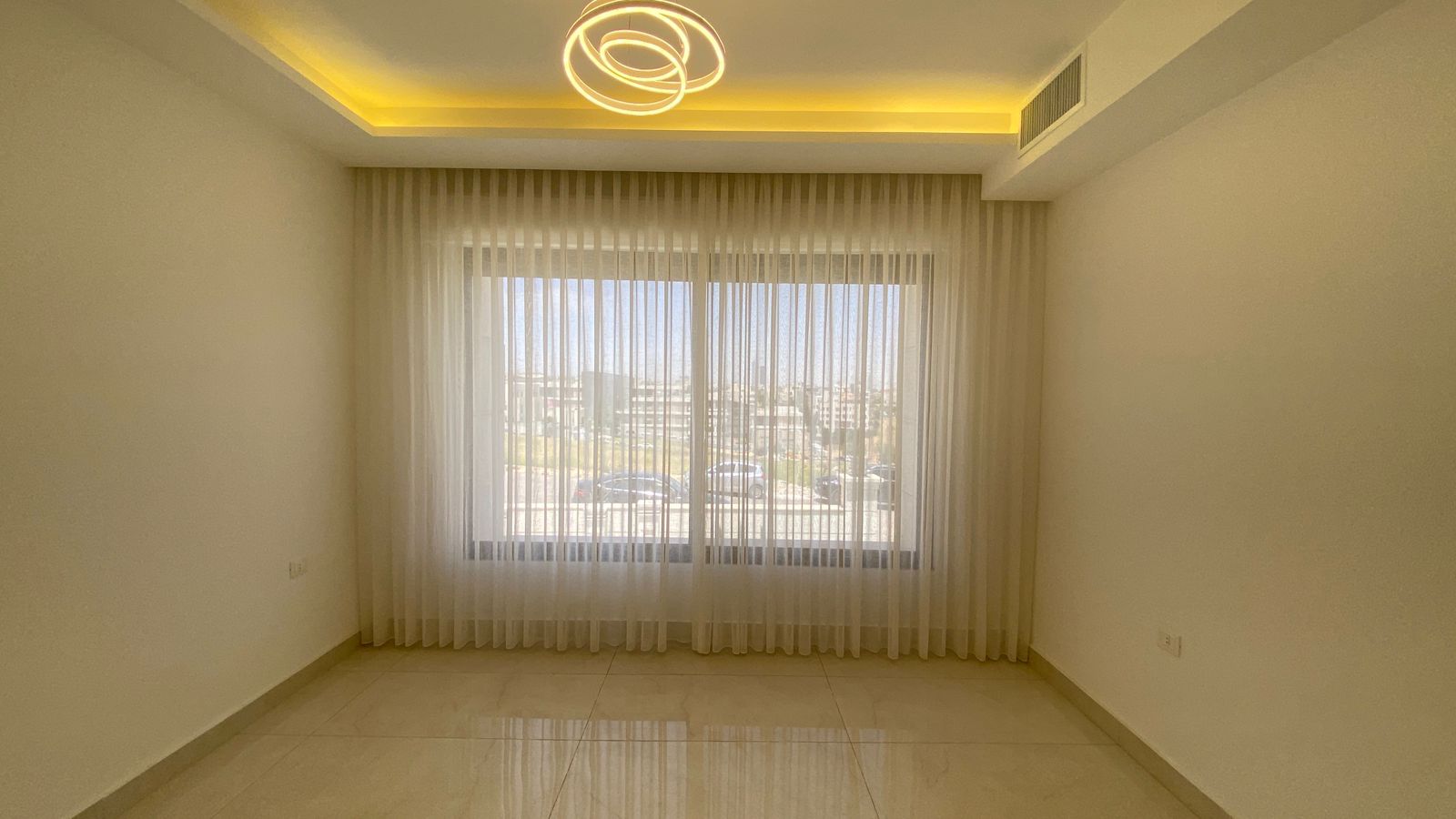 ca9ffc34-6186-4416-bc28-489a8554a967 Empty living room with a large window behind sheer white curtains, a glossy tile floor, and a circular ceiling light fixture.