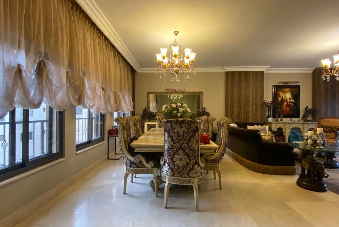 cb59ad46-1d6b-4e77-80cd-d39468bf789c Elegant dining room with ornate upholstered chairs around a long table, crystal chandelier overhead, and large windows with sheer drapes.