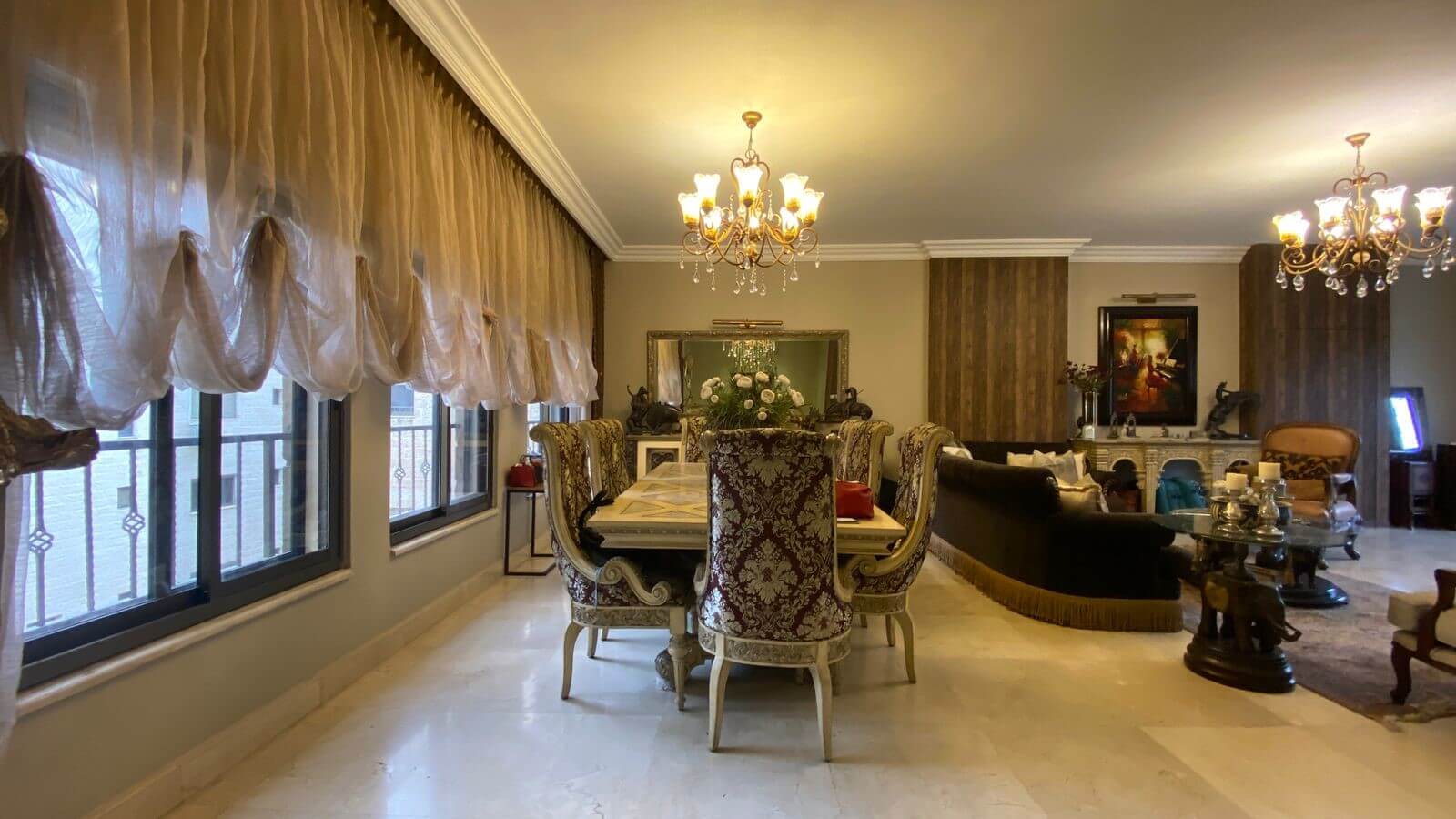 cb59ad46-1d6b-4e77-80cd-d39468bf789c Elegant dining room with ornate upholstered chairs around a long table, crystal chandelier overhead, and large windows with sheer drapes.