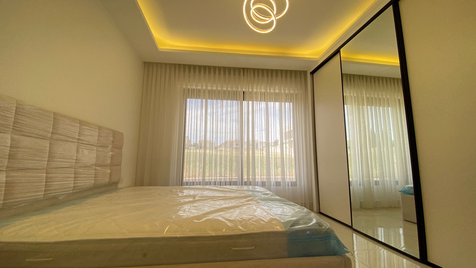 cc0342b6-d03a-43e3-a8c9-5c338fb13b93 Modern bedroom with a padded headboard, large window with sheer curtains, and a sliding mirrored wardrobe.