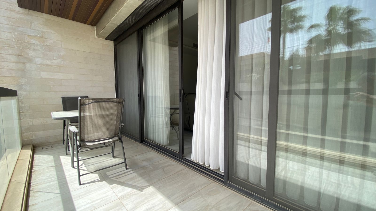 cc95a837-c450-475a-a2c2-8ff500362e1a Balcony with a small table and two chairs beside sliding glass doors and white curtains.