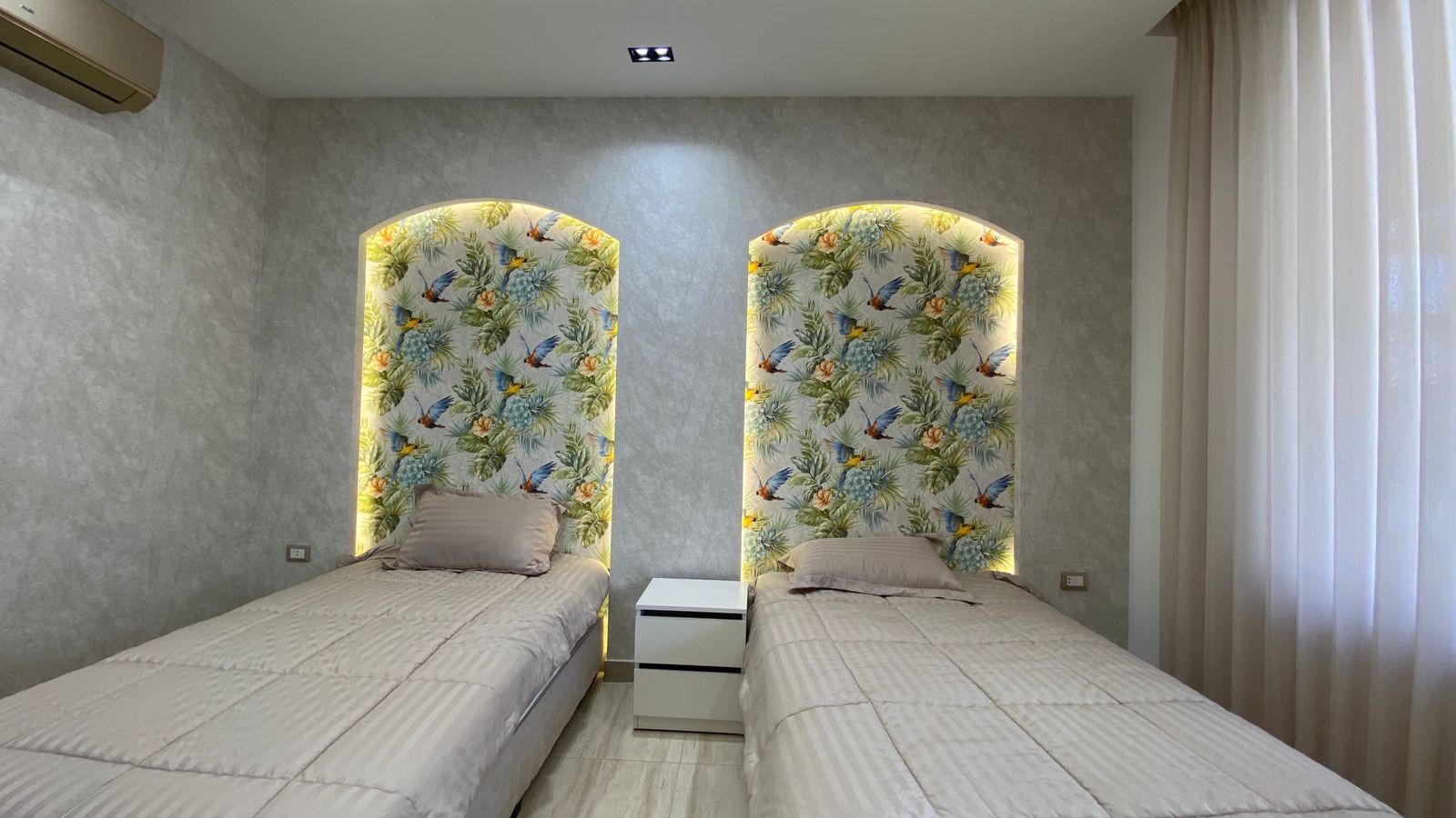 ccad1ca8-4b0b-4fe0-a88d-31f97d5e9126 Twin bedroom with tropical-patterned arched alcoves, each bed with beige pillows, a white two-drawer nightstand between them, and sheer curtains on the right side of the room.