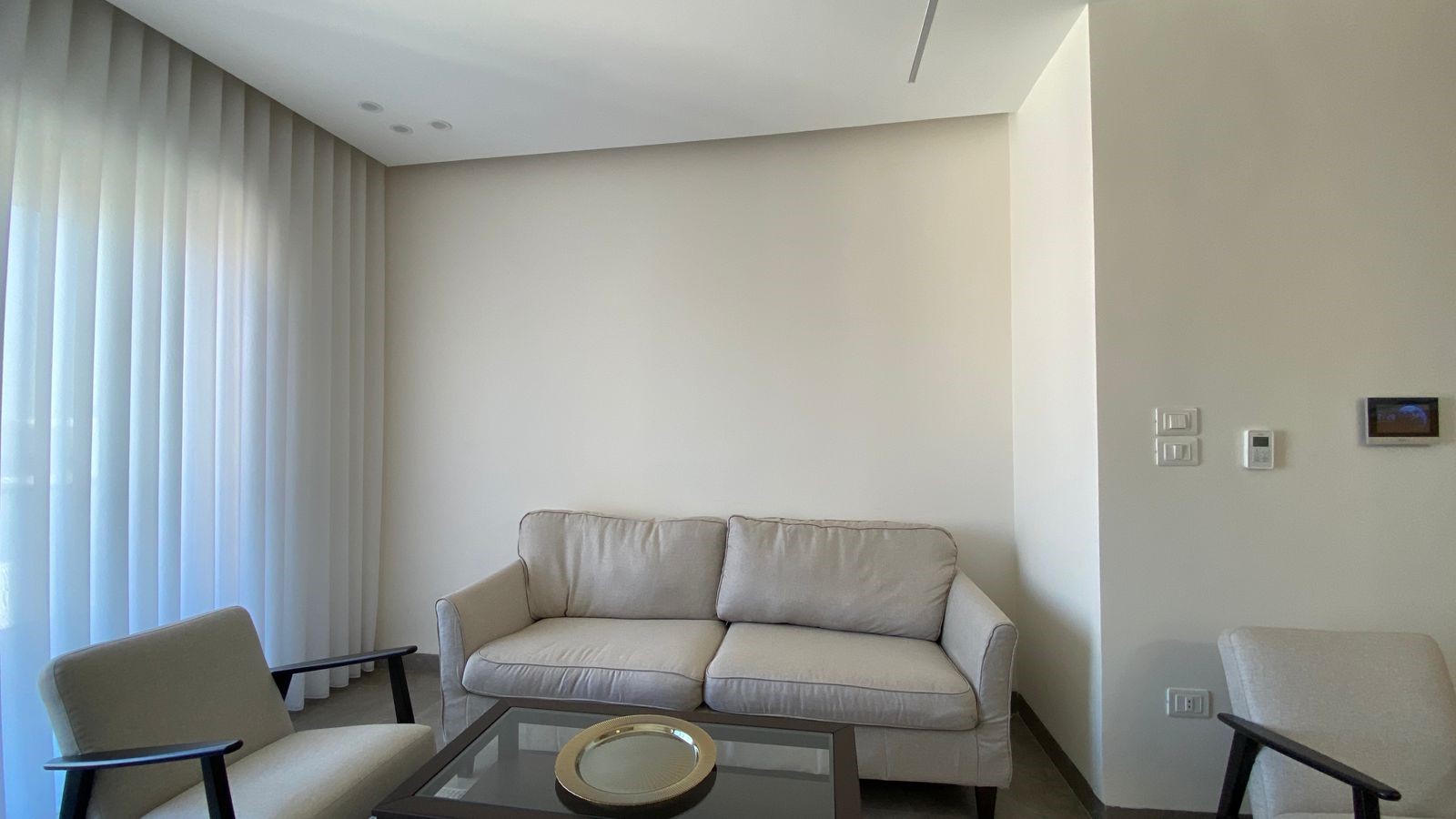 cedd08f4-2d90-4775-a96d-f8e86755f8aa Minimalist living room with a beige sofa and matching chairs around a glass coffee table, large sheer-curtained window on the left.