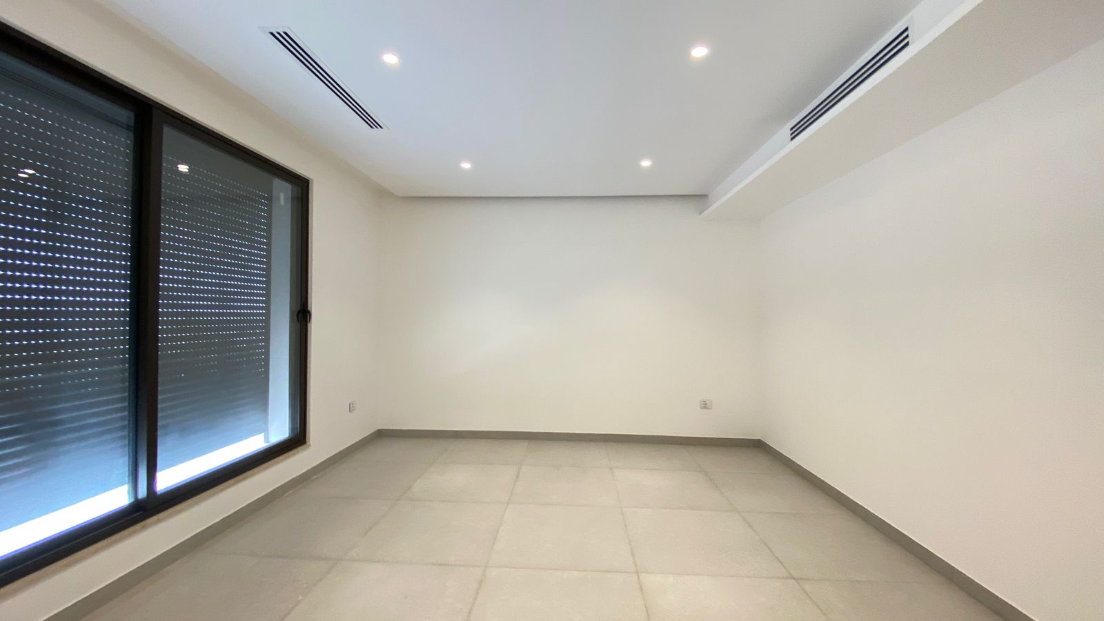 cf3657e7-f670-4cee-be0b-380470f45828 Empty modern room with a large sliding glass door and dark blinds, white walls, light gray tiled floor, and recessed ceiling lights with air vents above.