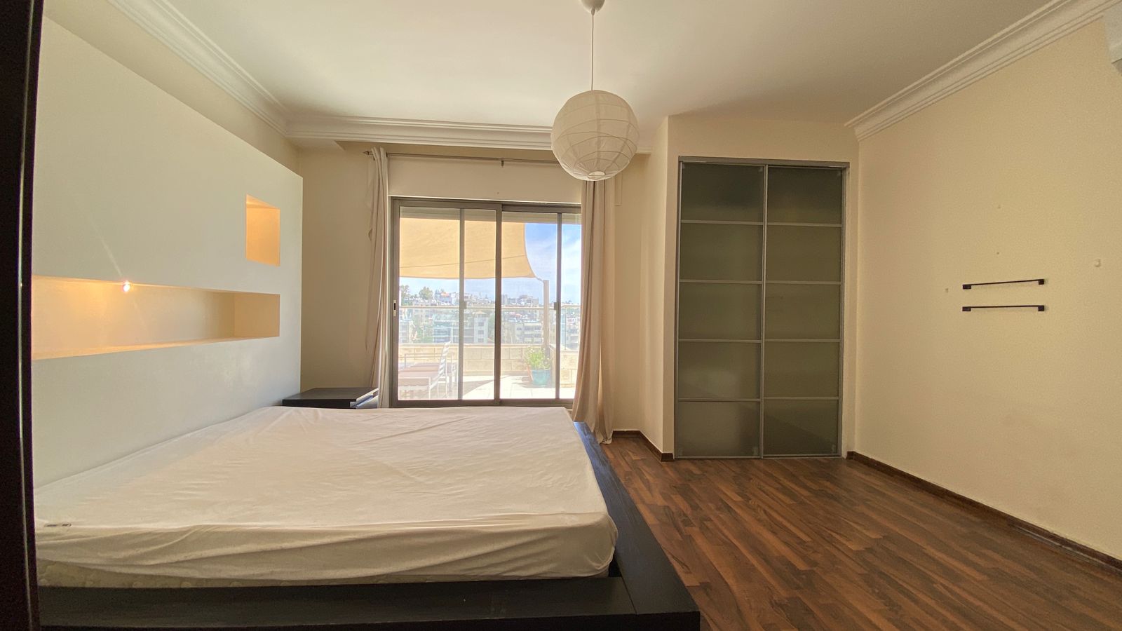 cf374a96-6060-4d2d-b137-a01a5d4e088b Bedroom with a low platform bed against a white headboard that has built-in shelves and a small light, sliding glass door to a balcony with city view, beige curtains, and a round paper lampshade overhead.