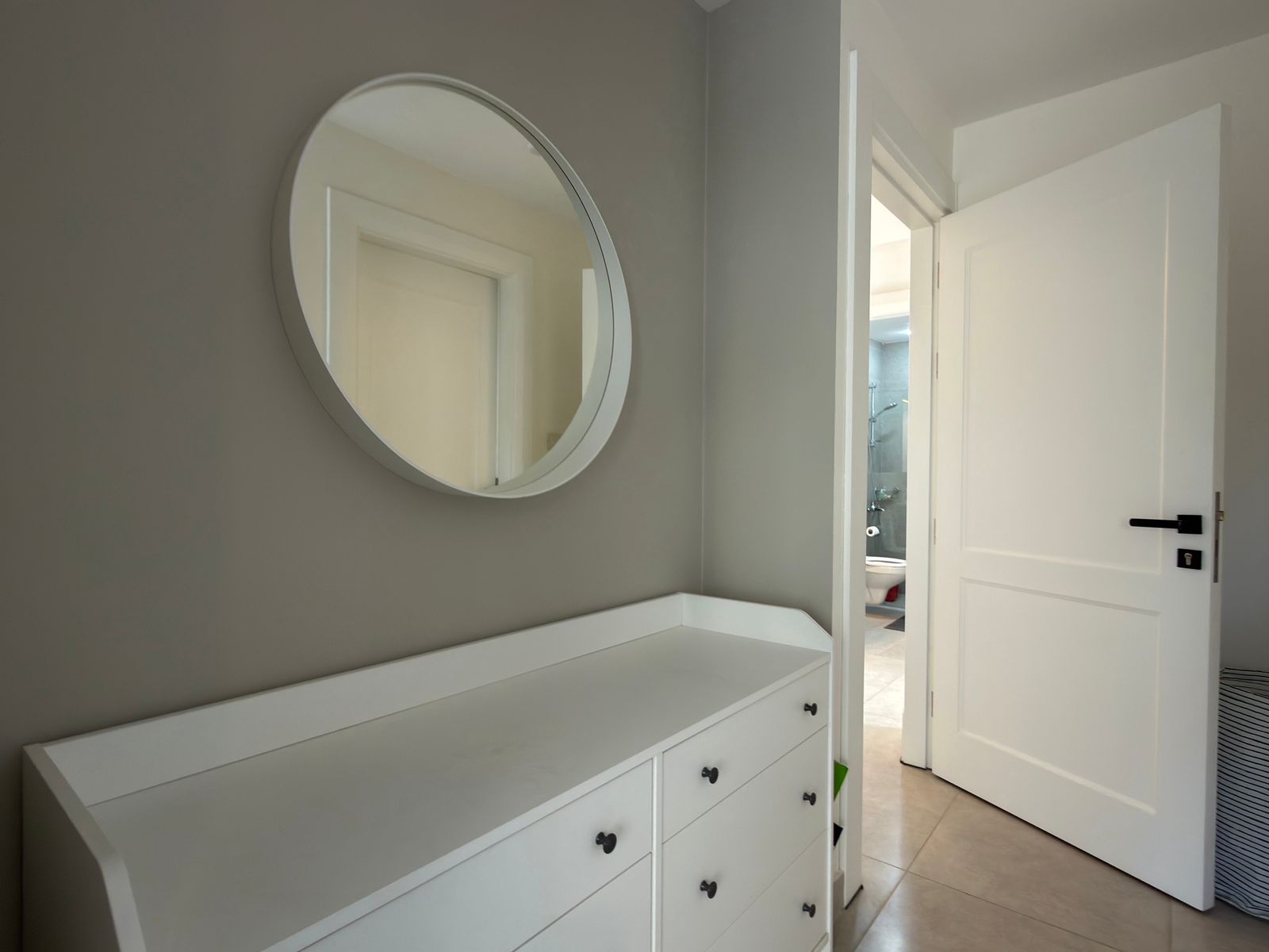 d041ed3d-0caa-41e4-932f-d0ef9d2446b9 White dresser with multiple drawers beneath a round wall mirror, in a gray-painted room; an open door reveals a bathroom with a glass shower enclosure beyond.