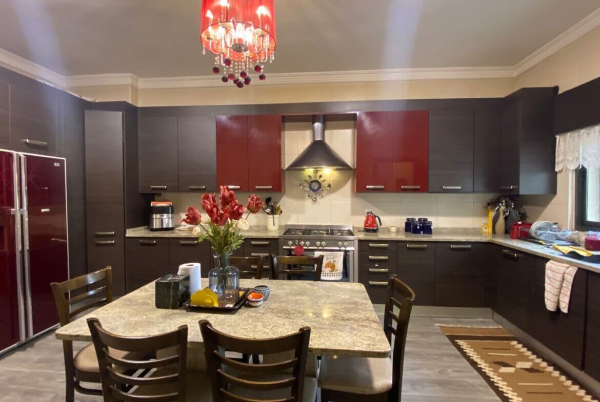 d0871463-cded-4d89-9d2c-9bbf0fd613bc Modern kitchen with dark cabinetry, red upper cabinet doors, and a stainless refrigerator surrounding a central island. A red chandelier hangs from the ceiling.