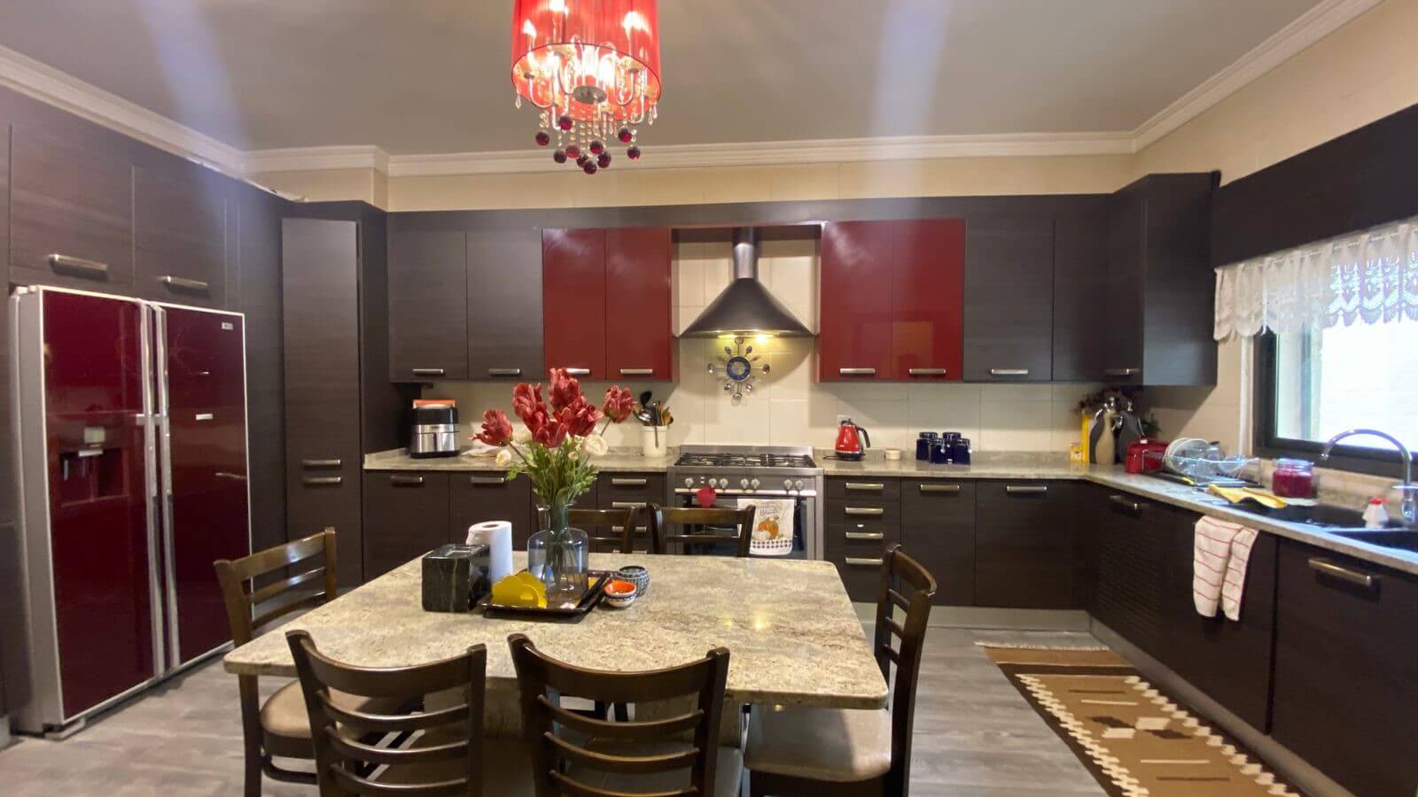 d0871463-cded-4d89-9d2c-9bbf0fd613bc Modern kitchen with dark cabinetry, red upper cabinet doors, and a stainless refrigerator surrounding a central island. A red chandelier hangs from the ceiling.