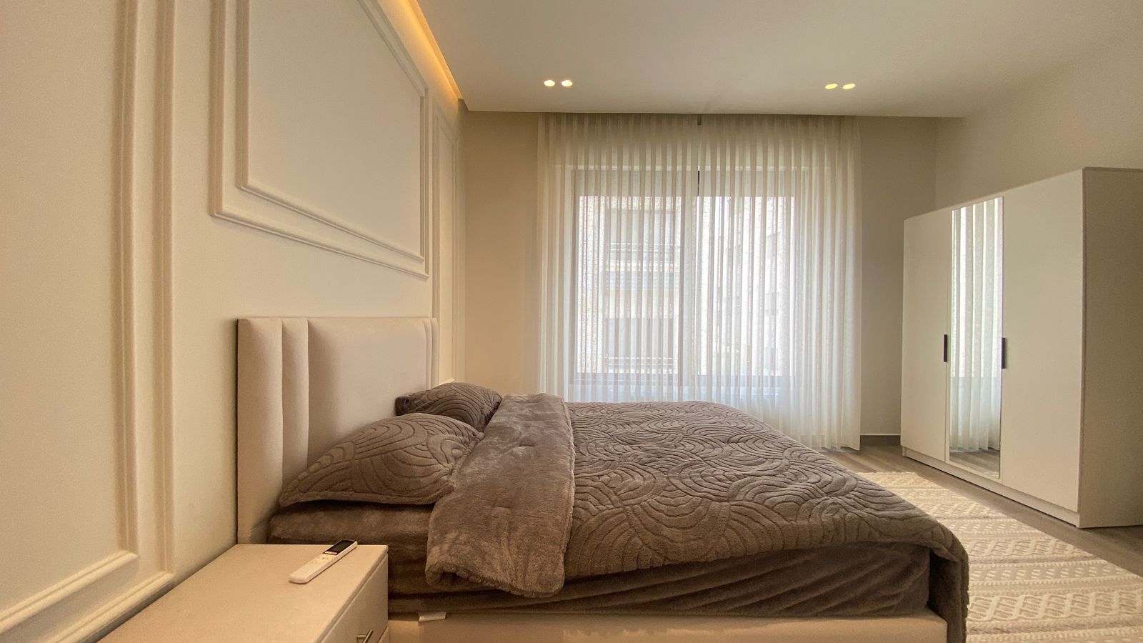 d0874ff4-e5bb-4ccd-bb67-6a85ea1f867a Modern bedroom with low, upholstered bed, gray quilt, and matching pillows; white nightstand with remote.