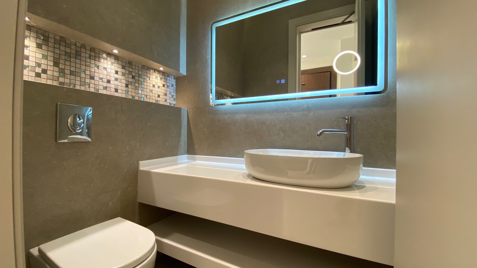 d0eec480-7a94-440a-9e9f-5e47001c4ce9 Modern bathroom with a wall-mounted white vessel sink, chrome faucet, backlit rectangular mirror, mosaic tile accent, and wall-hung toilet.