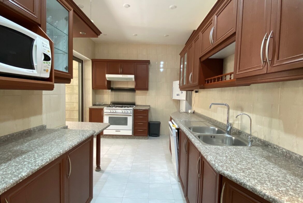 Bright galley kitchen with brown cabinets, granite counters, double sink, and a white oven against tiled walls in the back corner.