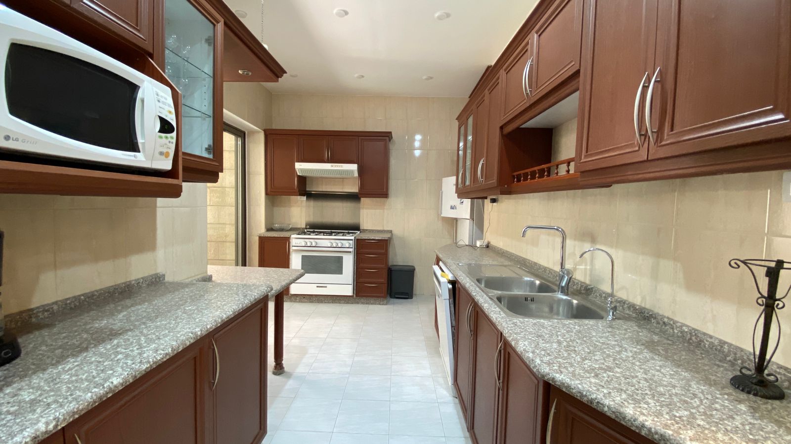 d2f55971-7014-4c61-a1a8-9a4b5bc2fecc Bright galley kitchen with brown cabinets, granite counters, double sink, and a white oven against tiled walls in the back corner.