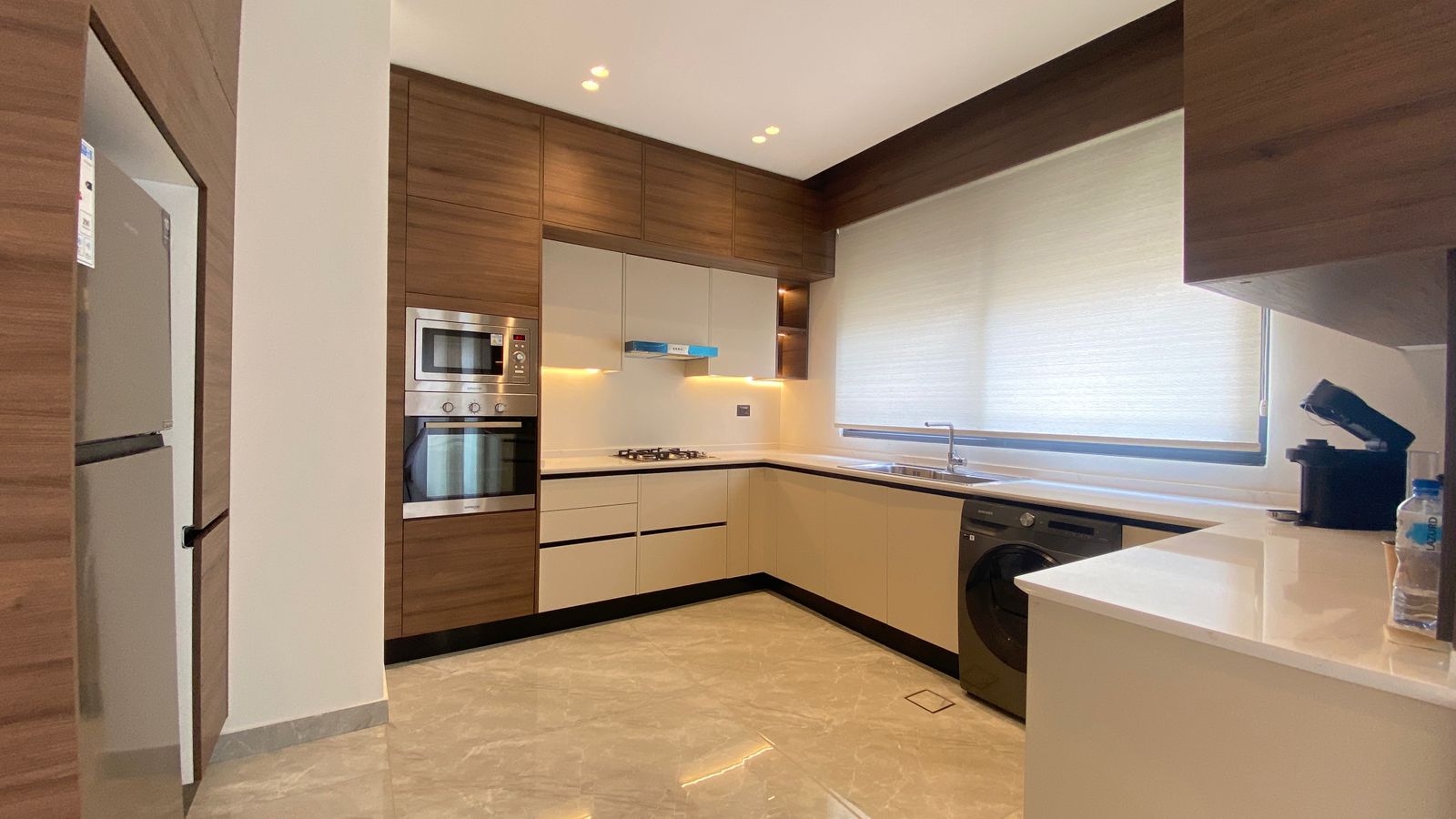 d3ee1937-49a4-48df-b774-3cfa251e4d2e Modern L-shaped kitchen with wood cabinets, white countertops, stainless steel oven and microwave, gas cooktop, and a front-loading washing machine beneath the counter.