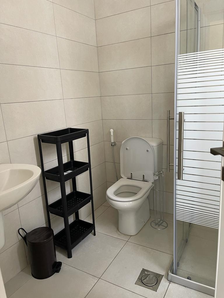 d45c26d5-b14a-4a38-81bf-89ef10593c7f Small modern bathroom with white toilet, glass shower, white sink, black open-shelf rack, and trash can in the corner.