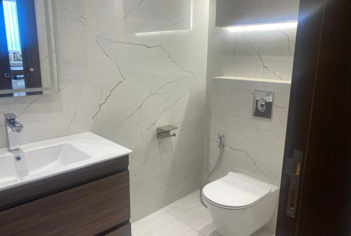 Modern bathroom with white marble walls, a wall-mounted toilet with chrome flush, a handheld bidet hose, and a dark wood vanity.