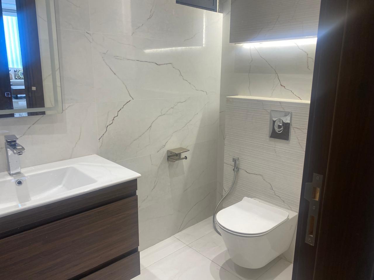 Modern bathroom with white marble walls, a wall-mounted toilet with chrome flush, a handheld bidet hose, and a dark wood vanity.