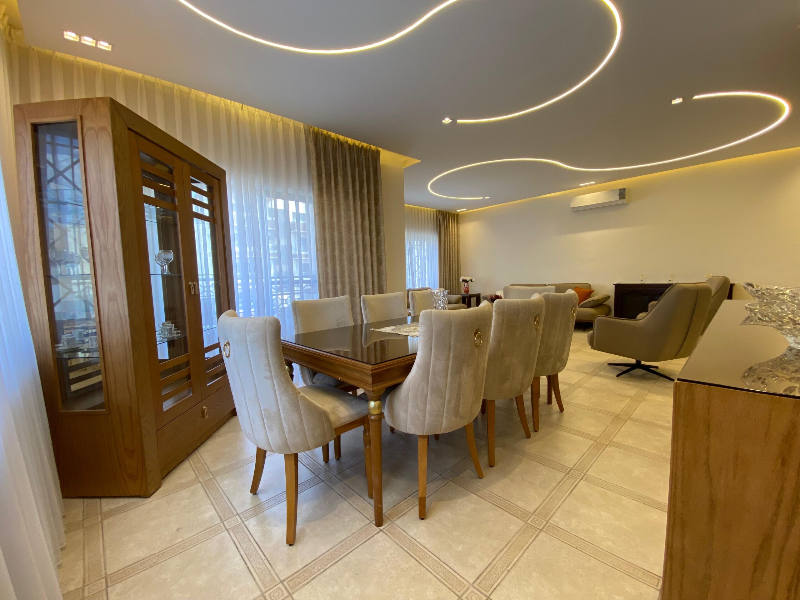 d56a4759-f77e-42dc-83cb-66379f367b9f Modern dining room with a long wooden table, beige upholstered chairs, and a glass-front cabinet near tall windows with sheer curtains.