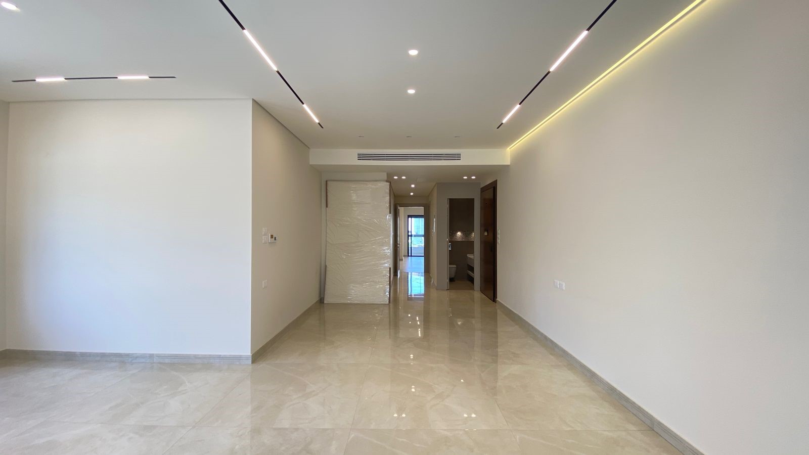 d5963ec0-f77b-49f1-a037-90b46d498533 Empty modern apartment hall with glossy tile floor, white walls, and linear ceiling lights; a large wrapped object rests against the left wall.
