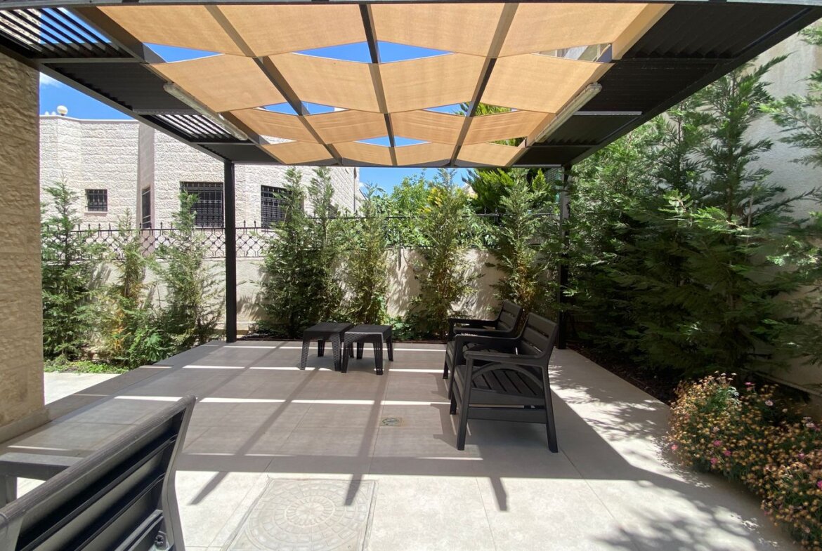 Outdoor seating area under a tan geometric shade sail, with black chairs and a small table, surrounded by tall evergreen hedges and flowering plants.