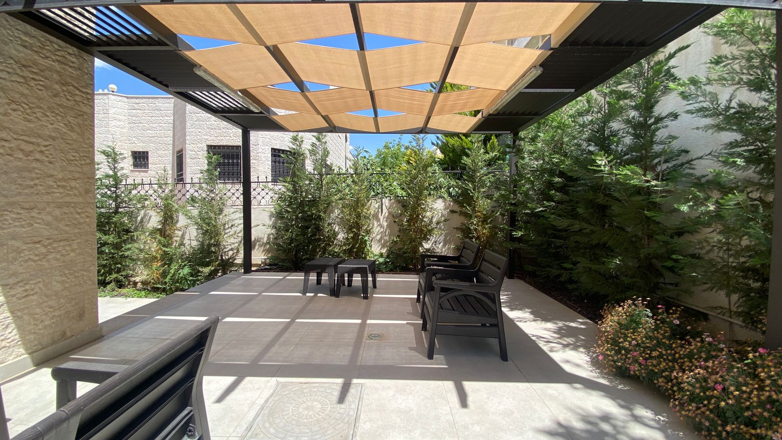 d5a78f59-7669-42d8-ad9c-4e6a5e7bf26a Outdoor seating area under a tan geometric shade sail, with black chairs and a small table, surrounded by tall evergreen hedges and flowering plants.