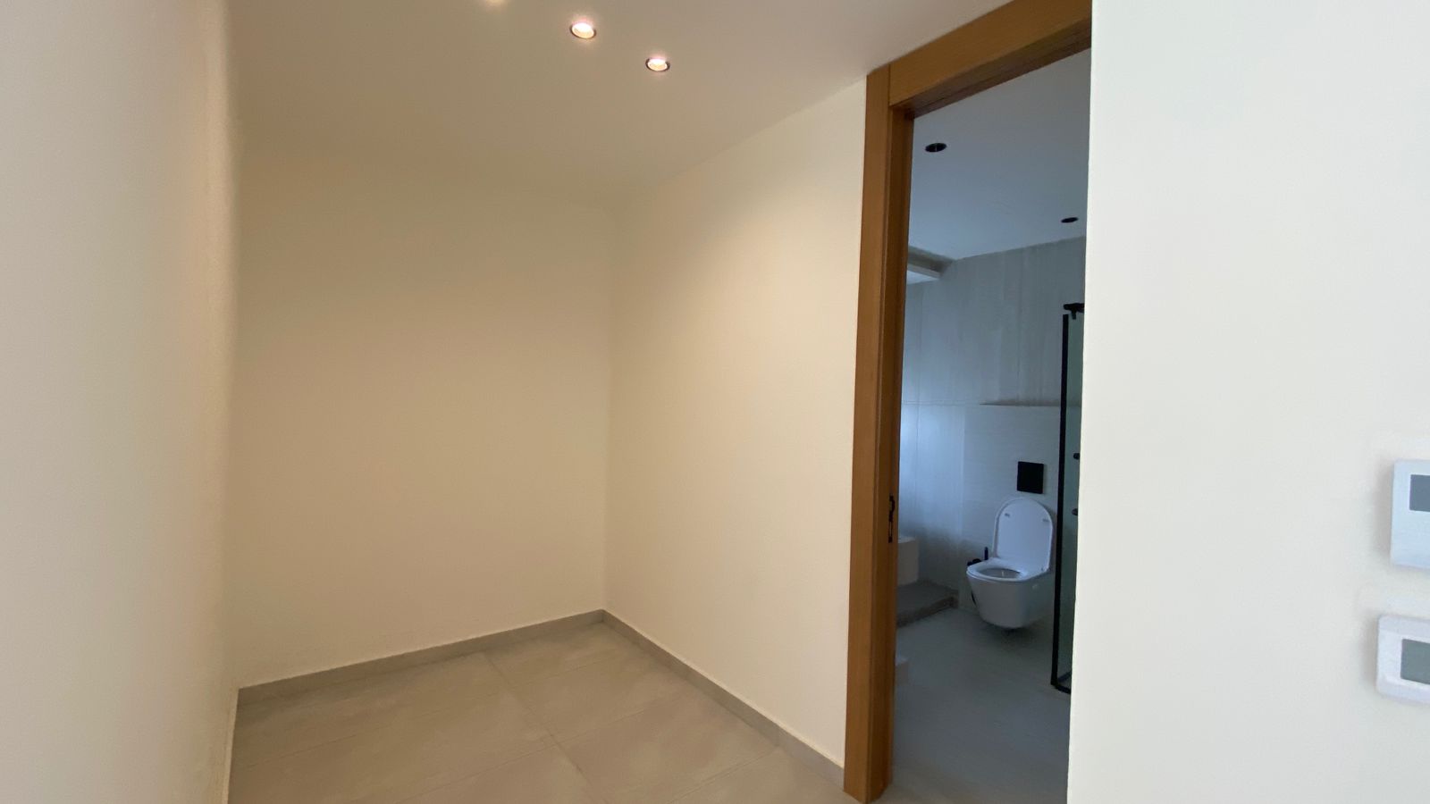d6407b48-72ca-4136-9c22-0e3b9b4827aa Empty hallway with beige walls and tiled floor; open wooden doorway reveals a bathroom with a toilet visible beyond.
