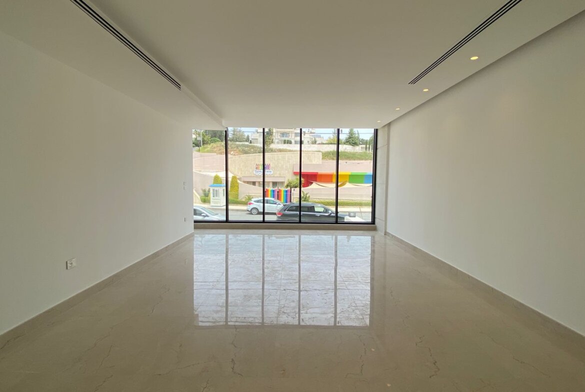 Empty modern showroom with glossy marble floor, white walls, recessed ceiling lights, and large front windows overlooking the street.
