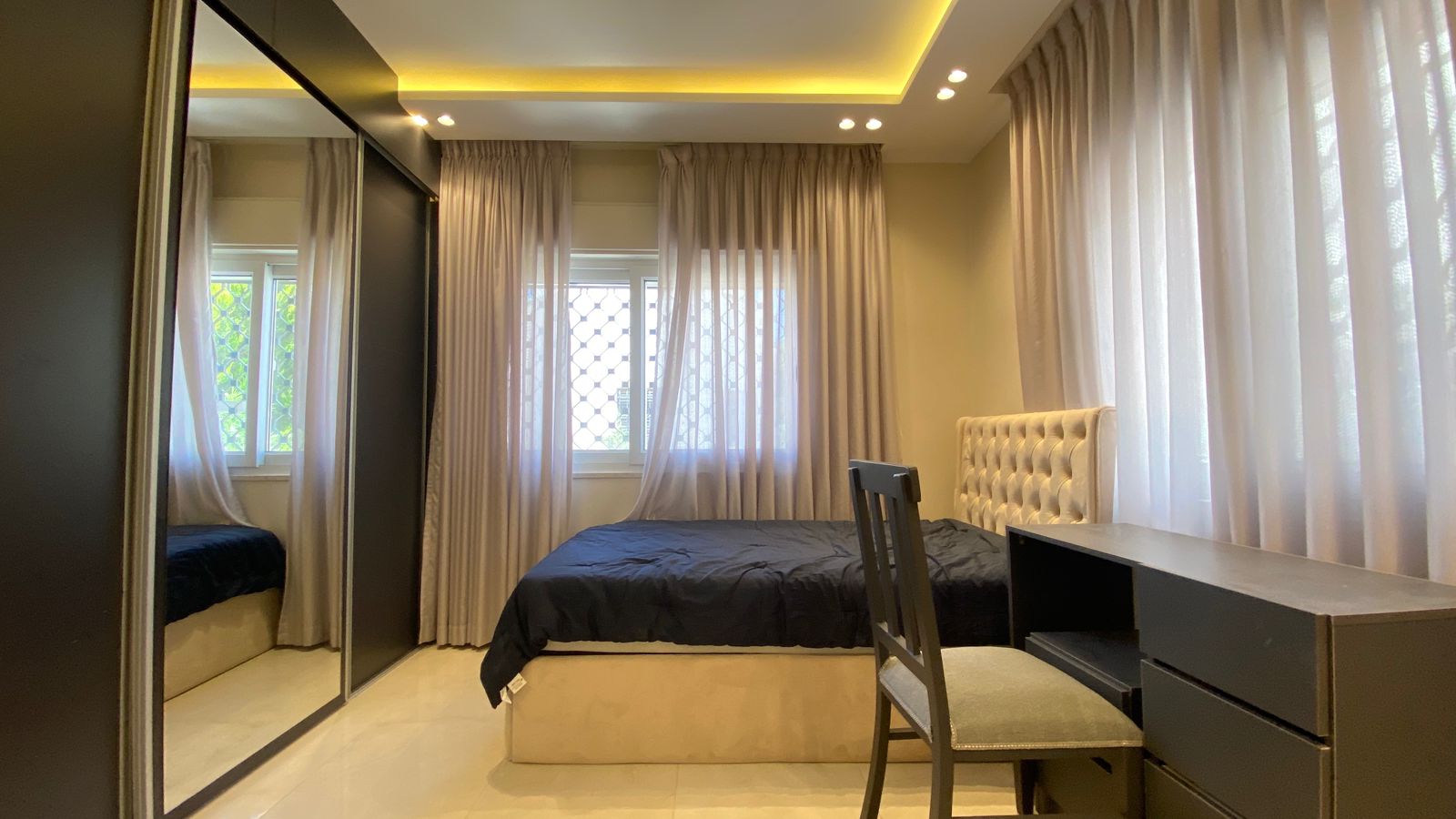d6c074ff-346a-4a07-b034-6aff4464d19f Bedroom with tufted beige headboard, dark bedspread, and large windows with sheer curtains in a sunlit room