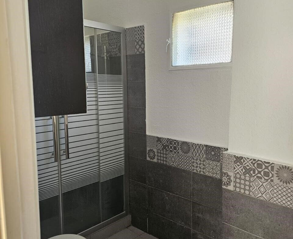 d6e0b12d-b072-4b32-92c0-b67d57c22f16 Small bathroom with a corner shower, toilet, and tiled walls featuring dark gray and decorative patterned tiles; high window on the right.