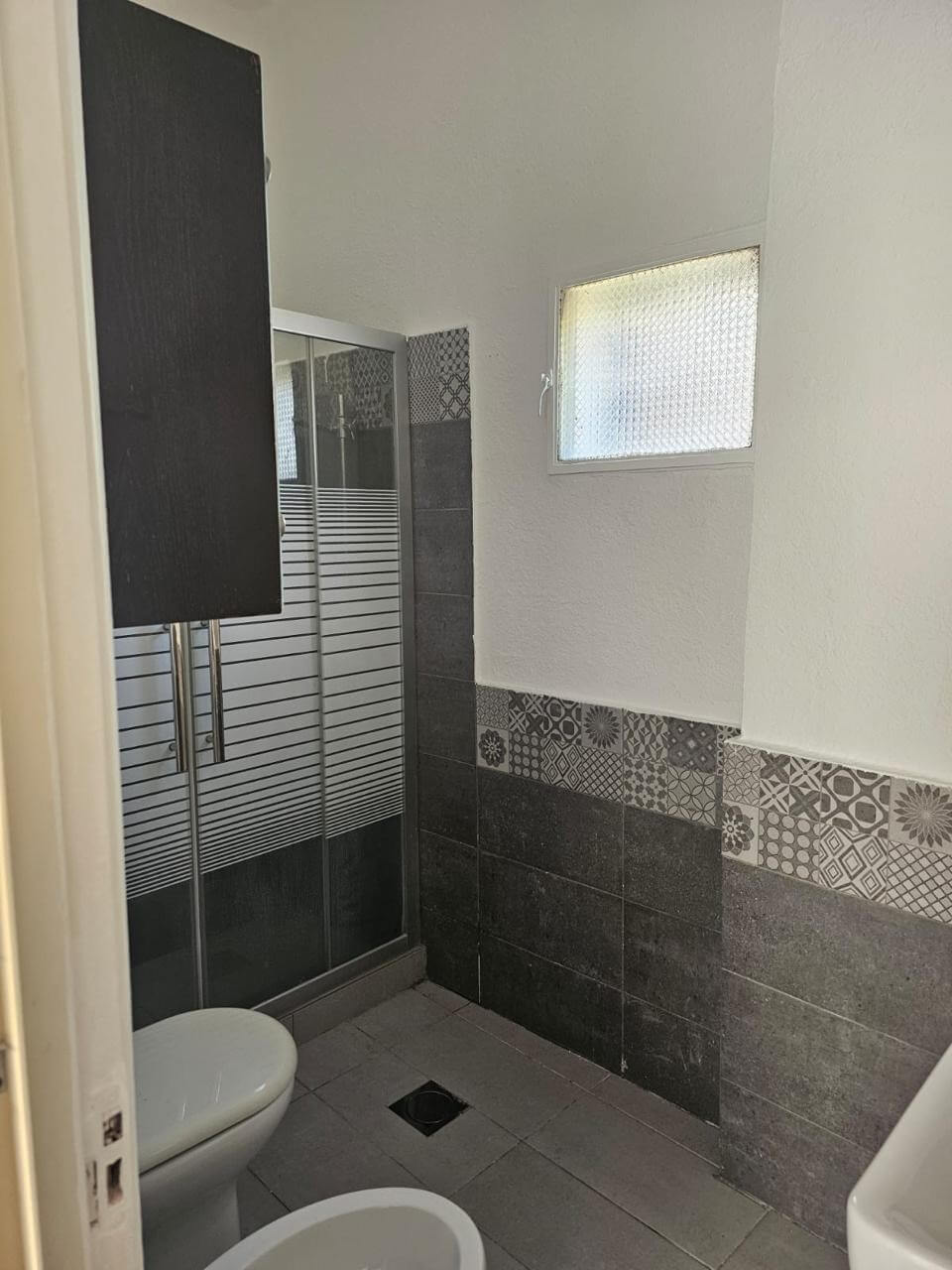 d6e0b12d-b072-4b32-92c0-b67d57c22f16 Small bathroom with a corner shower, toilet, and tiled walls featuring dark gray and decorative patterned tiles; high window on the right.